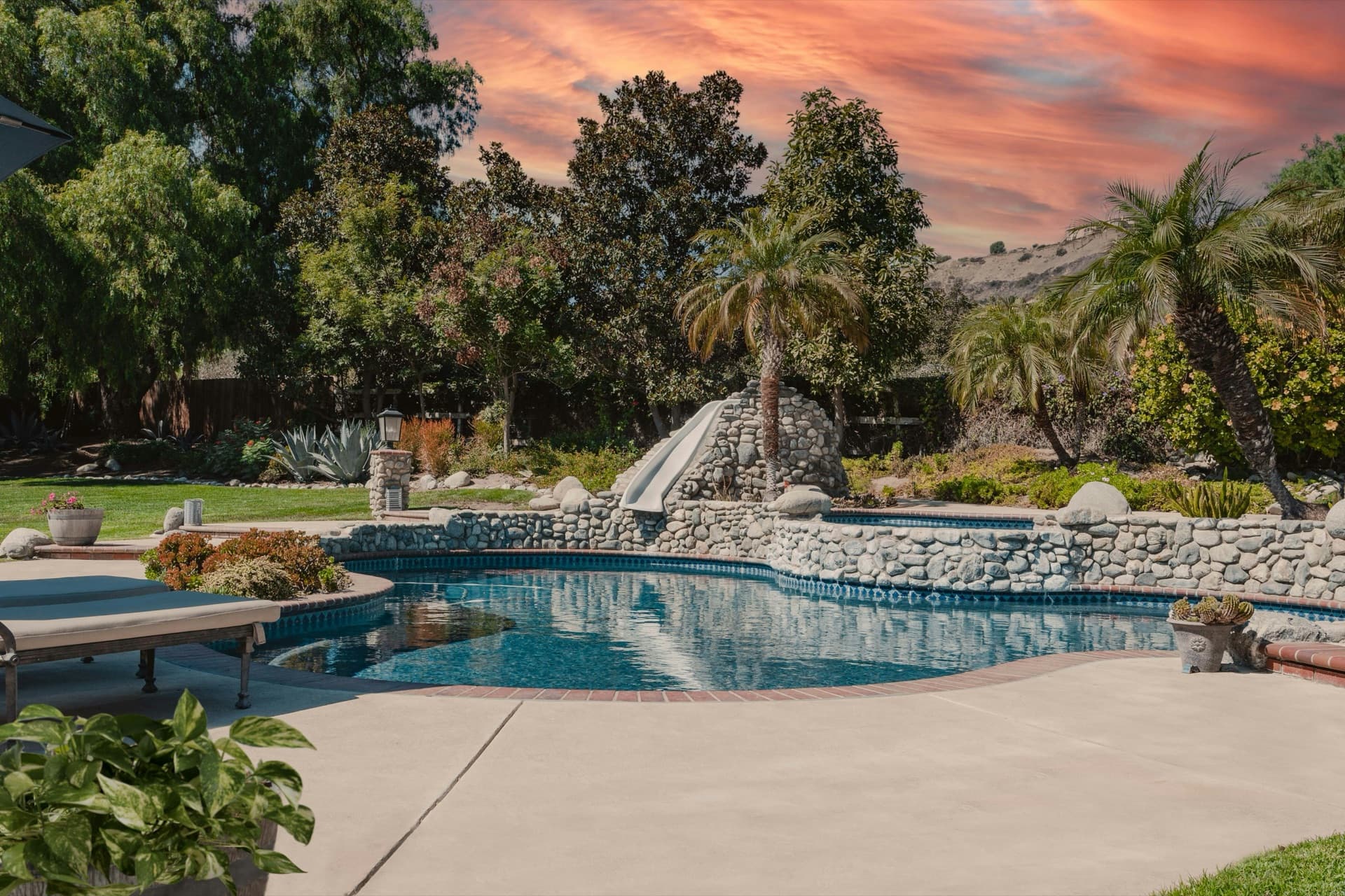 Backyard pool with a stone waterfall and slide, surrounded by landscaped gardens at sunset