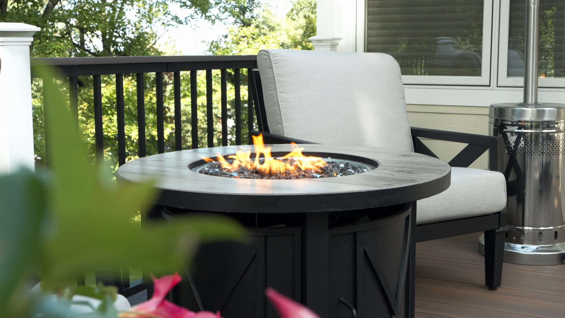 A gas fire pit burning on a backyard deck, framed by leaves in the foreground